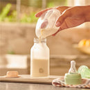 Silicone breastmilk collector cup pouring milk into a baby bottle, with a pacifier and storage container nearby.