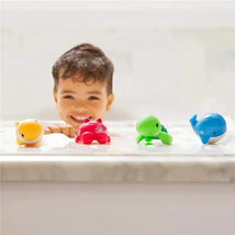 Four colorful ocean-themed bath squirters—a yellow fish, red crab, green turtle, and blue whale—are in a bubbly bathtub alongside a smiling child.