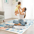 Mother joyfully holding a baby in a yellow onesie, seated on a colorful rug near a Munchkin Designer Diaper Changer.