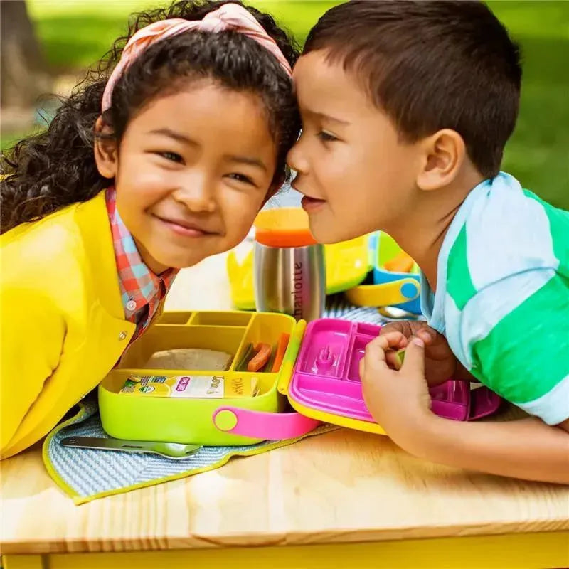 Two children interact playfully over a vibrant Munchkin Lunch Bento Box, featuring green and yellow colors, with stainless steel utensils visible.