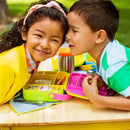 Two children interact playfully over a vibrant Munchkin Lunch Bento Box, featuring green and yellow colors, with stainless steel utensils visible.