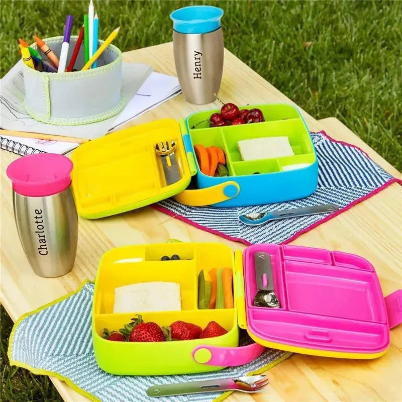 Bento boxes in green, yellow, and pink with stainless steel utensils; filled with fruits, vegetables, and sandwiches on a picnic table.