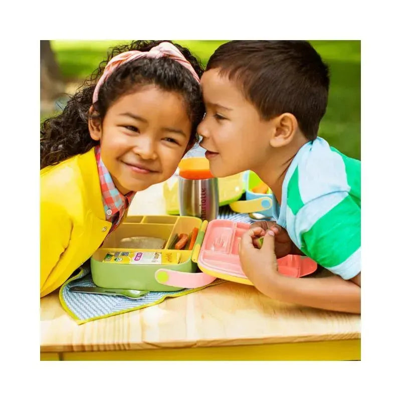 Two smiling children sit at a table with a Munchkin Lunch Bento Box featuring green and pink sections, along with stainless steel utensils.