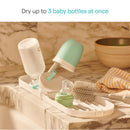 Munchkin Portable Drying Rack with space for three baby bottles, featuring a bottle brush and a striped towel on a marble countertop.