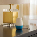 Portable white and blue sleep machine with control buttons, placed on a dark tabletop beside a lamp and decorative items.