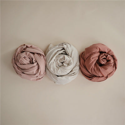 Three rolled baby wrap carriers in earthy tones: pink, beige, and cedar, on a light background.