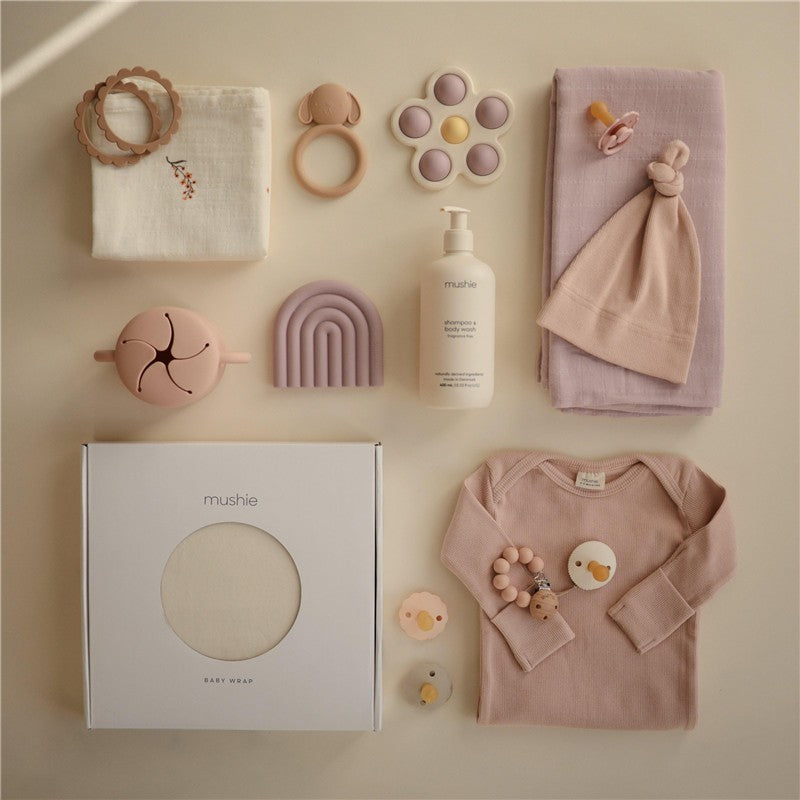Mushie 100% Organic Cotton Baby Wrap Carrier in Cedar, surrounded by a variety of baby accessories and clothing items.