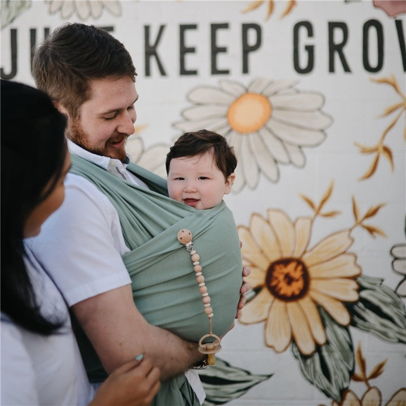 Mushie 100% Organic Cotton Baby Wrap Carrier in Roman Green, with a smiling baby held by a man, against a floral mural background.