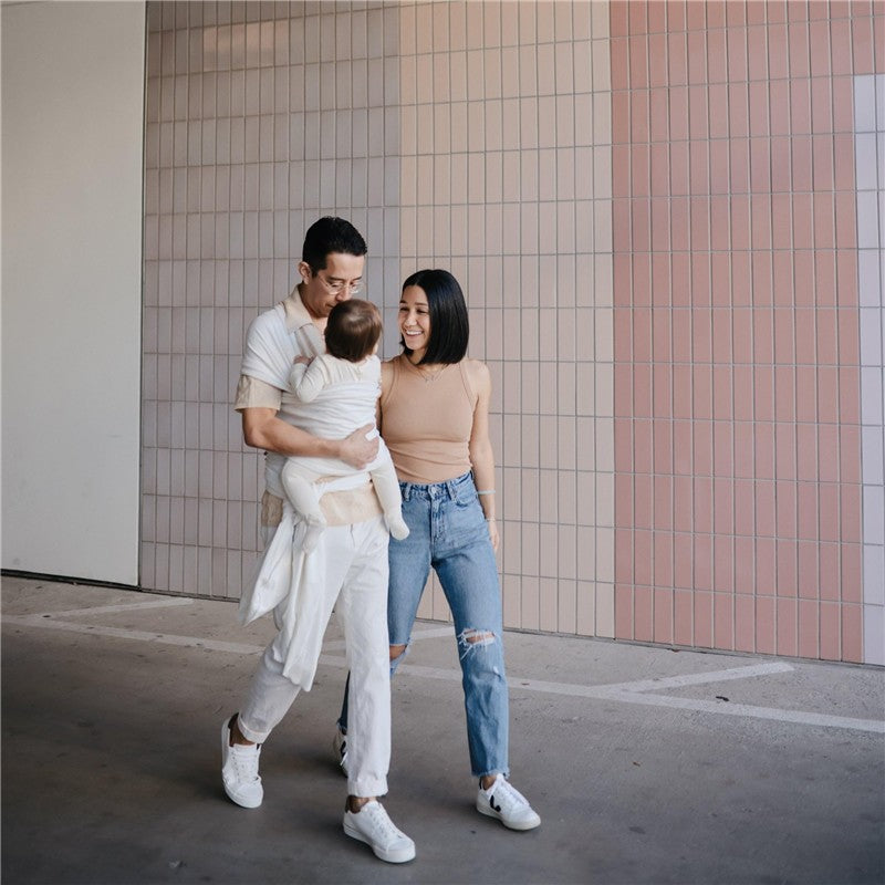 Couple smiles while walking in a pastel-colored area, one parent carries a baby wrap carrier with a child.