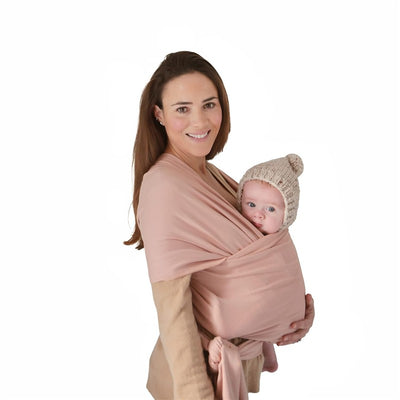 A woman holds a baby securely wrapped in a blush-colored organic cotton carrier, both smiling. The baby wears a cozy knit hat.