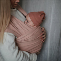 Mushie 100% organic cotton baby wrap carrier in blush color with a parent holding a baby wearing a matching hat.