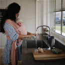 A woman in a blush organic cotton baby wrap is pouring water from a small cup in a kitchen sink while holding her baby.