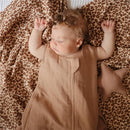 Baby wearing a natural-colored Mushie 100% Organic Cotton Muslin wearable blanket, surrounded by a leopard-patterned fabric and star-shaped toy.