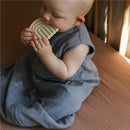 Baby wearing a blue organic cotton muslin blanket, holding a beige silicone rainbow teether while sitting on a bed.