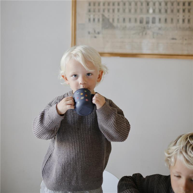 Toddler drinking from a Mushie silicone training cup with a planet design, wearing a cozy brown sweater.
