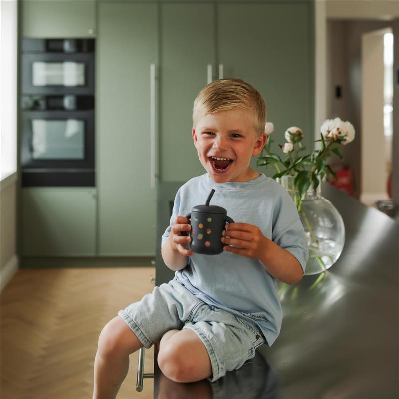 Toddler joyfully holding a Mushie silicone training cup with a straw, featuring bright dots, in a modern kitchen setting.