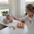 Mushie Baby Food Freezer Tray in Blush, with a mother feeding her baby on a high chair in a bright room.