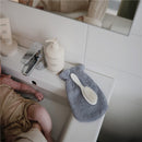 Mushie Organic Cotton Bath Mitts 2-Pack in gray on a bathroom sink, alongside baby cream and a brush.