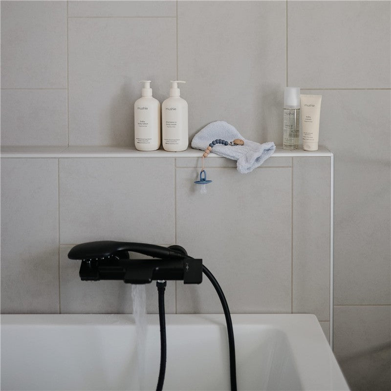 Gray organic cotton bath mitts from Mushie, displayed beside multiple bath products on a bath ledge.