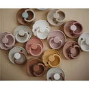 Mushie Frigg Daisy natural rubber pacifiers in various pastel colors, designed for infants 0-6 months, arranged on a surface.