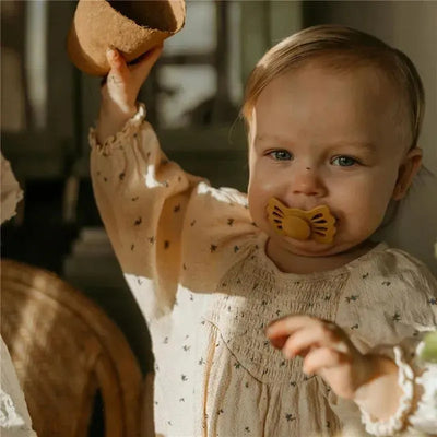 Toddler wearing a cream-colored blouse holding a small pot, smiling with a yellow silicone pacifier in their mouth.