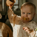 Toddler wearing a cream-colored blouse holding a small pot, smiling with a yellow silicone pacifier in their mouth.