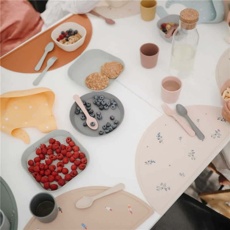 Mushie Silicone Round Plate Dinnerware Set in powder blue, featuring plates with fresh fruits and snacks on a table.