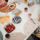 Mushie Silicone Round Plate Dinnerware Set in powder blue, featuring plates with fresh fruits and snacks on a table.