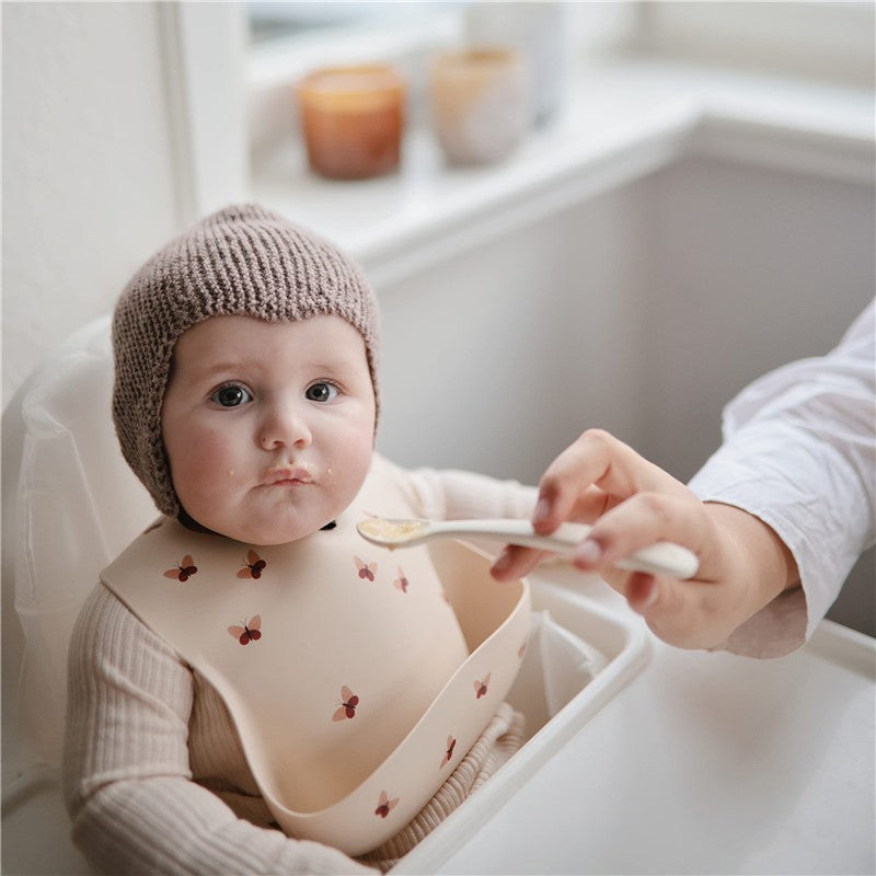 A baby in a high chair, wearing a beige bib with a red flower pattern, is being fed with a silicone spoon.