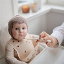 A baby in a high chair, wearing a beige bib with a red flower pattern, is being fed with a silicone spoon.