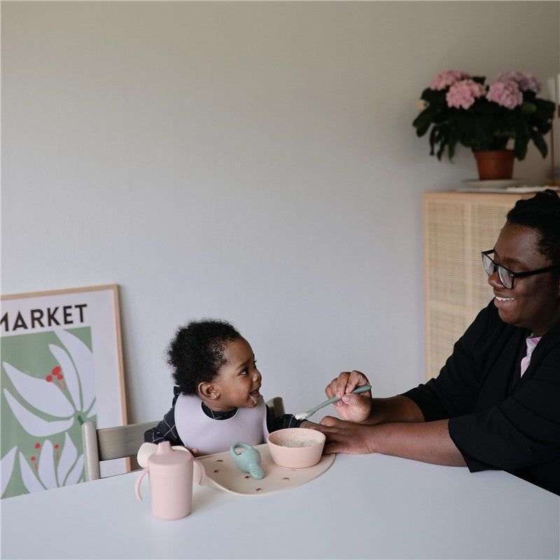 Mushie silicone feeding spoons in blush and shifting sand being used by an adult to feed a smiling child in a dining setting.