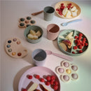 Mushie silicone feeding spoons in blush and shifting sand alongside colorful silicone plates and cups with different fruits.