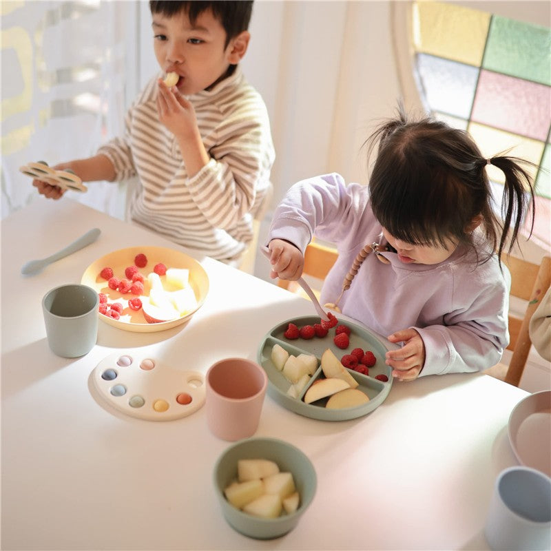 Children enjoying fruit on a table with Mushie silicone feeding spoons in Cambridge Blue and Shifting Sand colors.