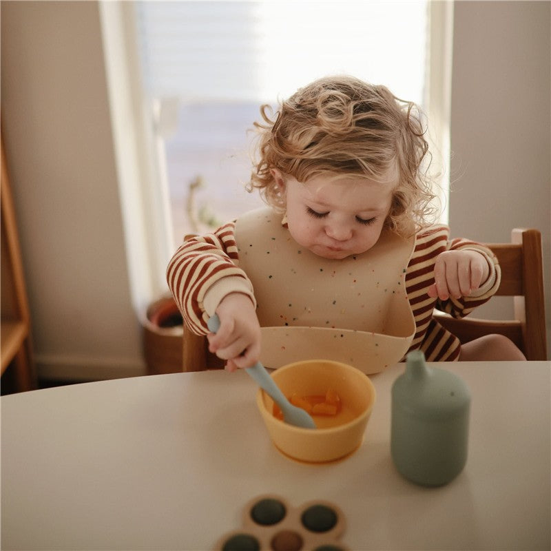 Child wearing a striped shirt and silicone bib, using a blue feeding spoon to eat from a yellow bowl.