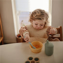 Child wearing a striped shirt and silicone bib, using a blue feeding spoon to eat from a yellow bowl.