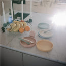 Mushie silicone feeding spoons in Cambridge Blue and Shifting Sand beside colorful plates and a fruit bowl on a marble surface.