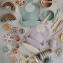 Mushie Silicone Feeding Spoons 2 Pack in Dried Thyme & Natural, with various colorful silicone kitchenware and utensils displayed.