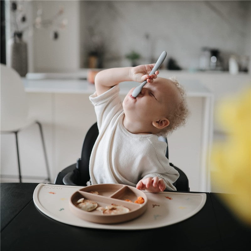 Toddler using a Mushie silicone feeding spoon, reaching for food on a divided plate while seated in a high chair.