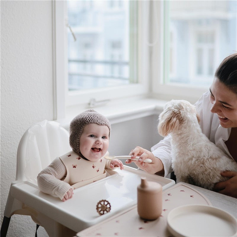 Mushie silicone feeding spoons in stone and cloudy mauve, used by a smiling baby in a high chair, with a mother and dog nearby.