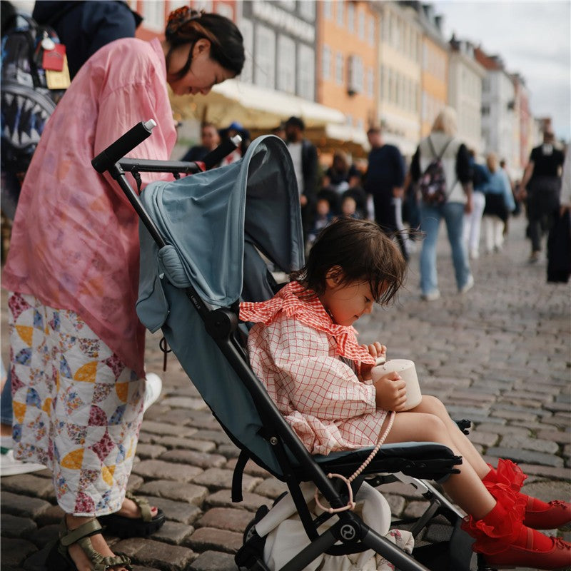 A child sitting in a blue stroller on cobblestone streets, holding a white cup, while an adult leans over nearby.