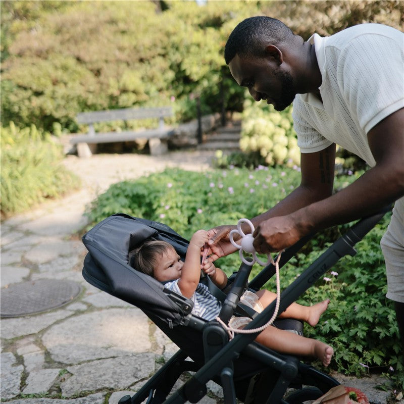 Father interacts with a baby in a stroller in a garden, adjusting a silicone strap with playful engagement.