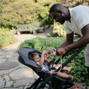 Father interacts with a baby in a stroller in a garden, adjusting a silicone strap with playful engagement.