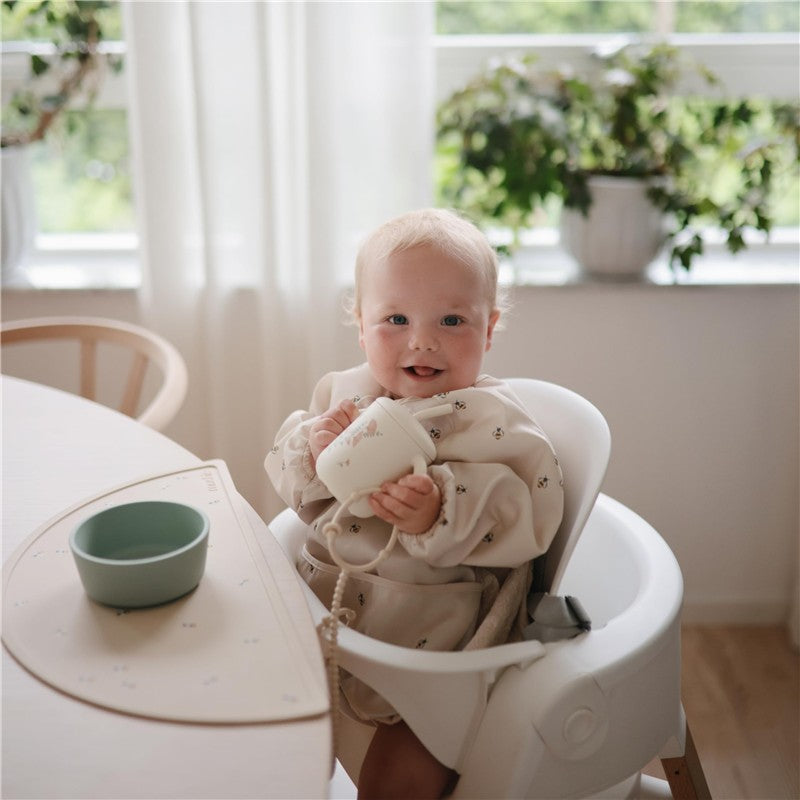 A happy child in a high chair holds a cup, with a green bowl on the table and plants in the background.