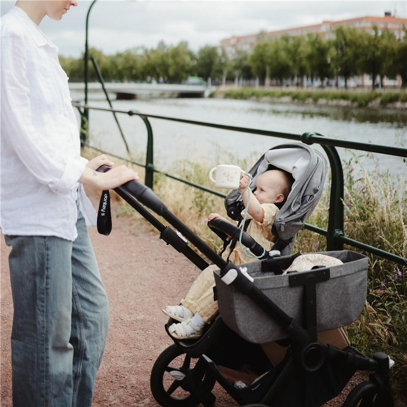 Mushie Adjustable Silicone Strap 2-Pack displayed with a parent pushing a stroller, containing a baby holding a cup.