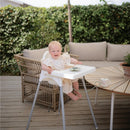 A child sitting in a high chair on a wooden deck, smiling, with a green bush backdrop and a table beside a plant.