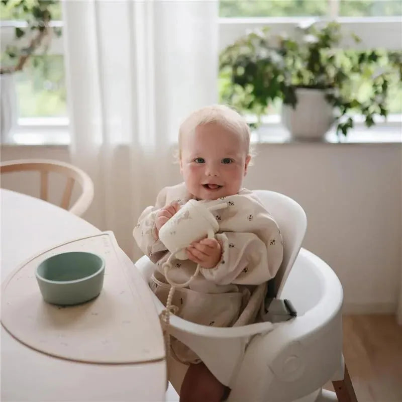 A smiling baby in a high chair holds a small cup, with a light-colored bowl on the table and plants in the background.