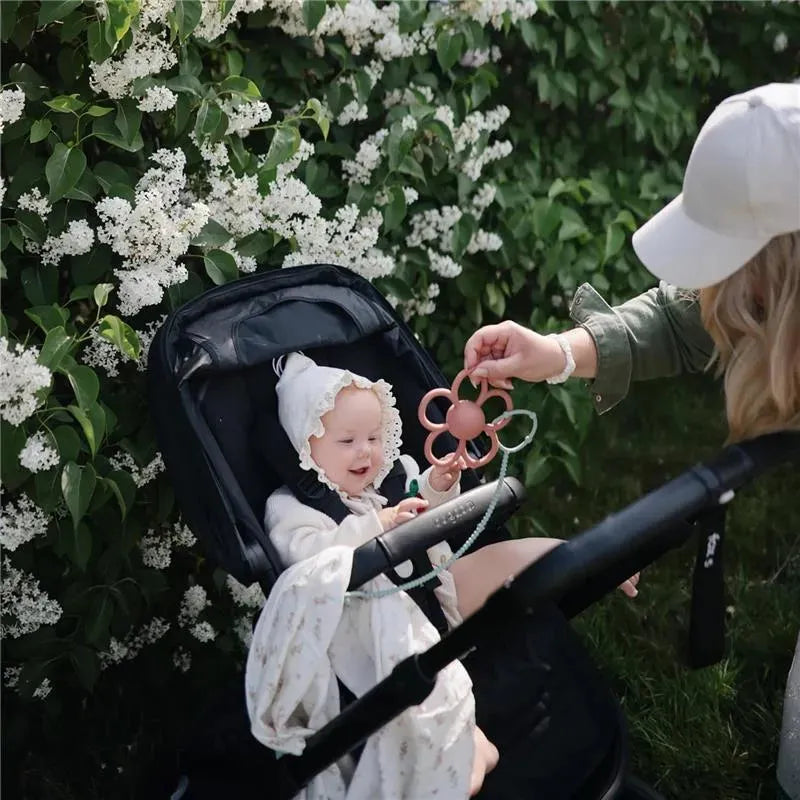 A smiling baby in a stroller, wearing a white bonnet, reaches for a pink flower-shaped toy held by an adult. Lush white flowers background.