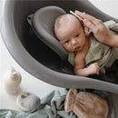 A baby reclines in a gray bath seat, being brushed with a comb while wrapped in a light green cloth, surrounded by bath products.