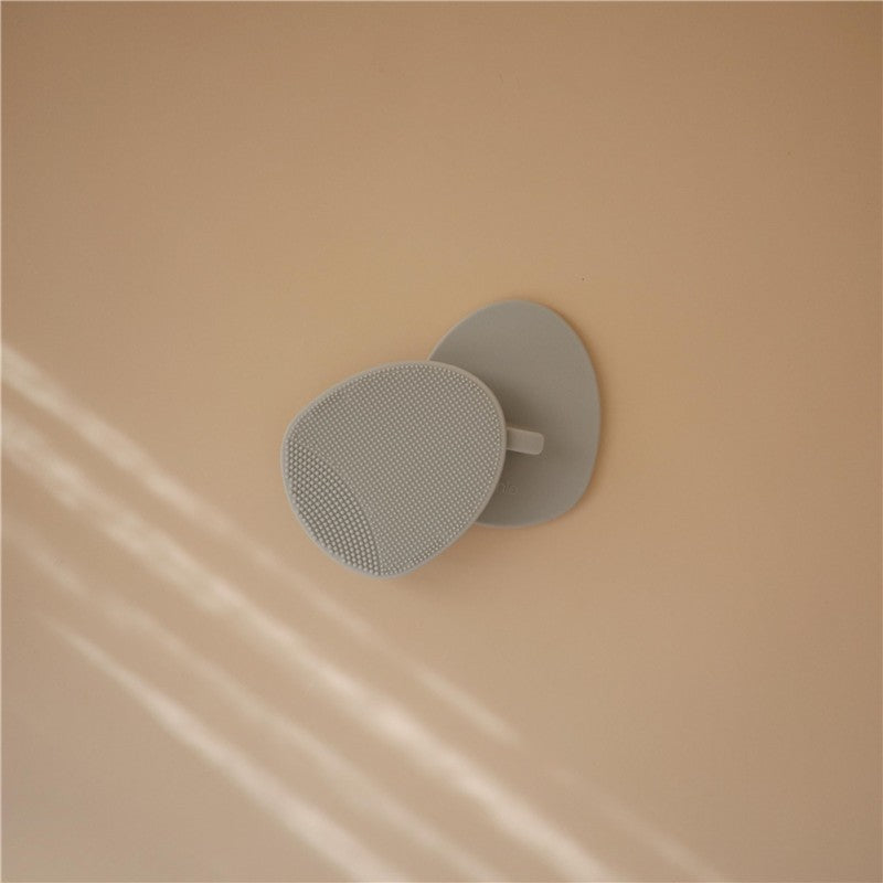Silicone baby bath cradle cap brush in light gray, featuring a textured surface and handle, resting against a peach background.