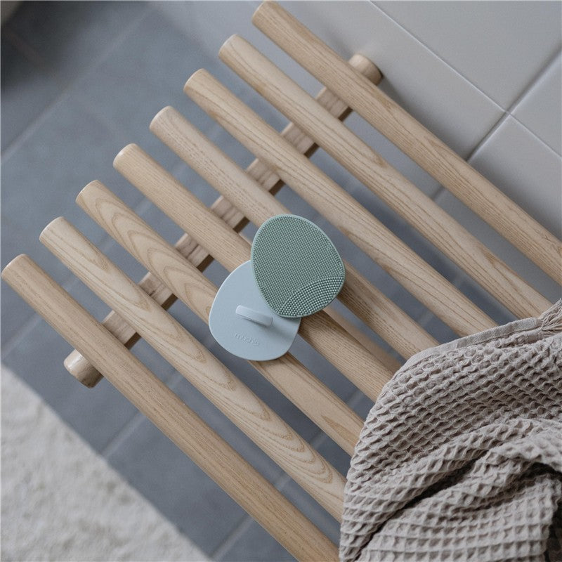 Mushie Silicone Baby Bath Cradle Cap Brush set in Tradewinds and Periwinkle, resting on a wooden rack beside a beige towel.